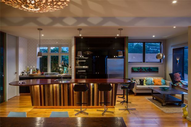 Open-plan kitchen and living area with a striking timber island featuring vertical striped wood detailing and a dark polished benchtop. The kitchen includes white brick-look wall tiling, dark glossy cabinetry, integrated appliances, pendant lighting and timber flooring. A contemporary living zone with a sofa, coffee table and large windows sits beyond the island. Warm, dramatic evening lighting and sculptural ceiling fixtures create a refined, inviting atmosphere.
