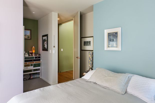 A cozy bedroom viewed from beside the bed, with a white bedspread and pillows in the foreground. The room has a muted blue feature wall, white trim and doors, warm timber flooring in the adjoining hallway, and grey carpet near a dark built-in dresser. Framed artwork and a small table lamp add understated decorative detail. An open doorway leads to a hallway or adjacent space.