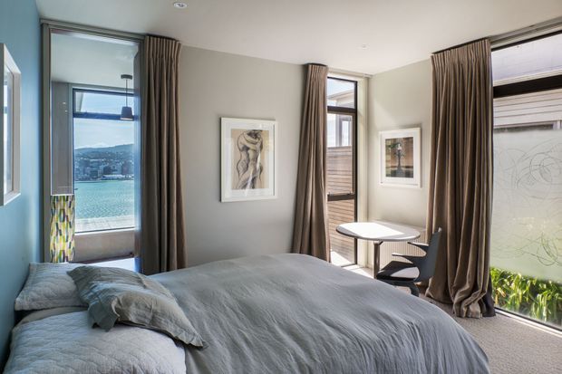 A bright contemporary bedroom with a low bed dressed in pale grey bedding, tall windows, and floor-to-ceiling taupe curtains. The room has grey painted walls, framed artwork, a small round table with dark chairs, and views toward a waterfront or hilly urban landscape through the glazing. Natural light creates an airy, calm atmosphere.