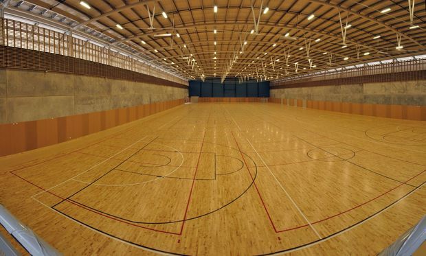A large indoor sports hall with a polished timber court marked for basketball and other court sports. The expansive space has tall exposed timber roof trusses, linear suspended lighting, high acoustic wall panels, and protective lower wall cladding. The view is wide-angle and symmetrical, emphasizing the scale of the multi-use gymnasium.
