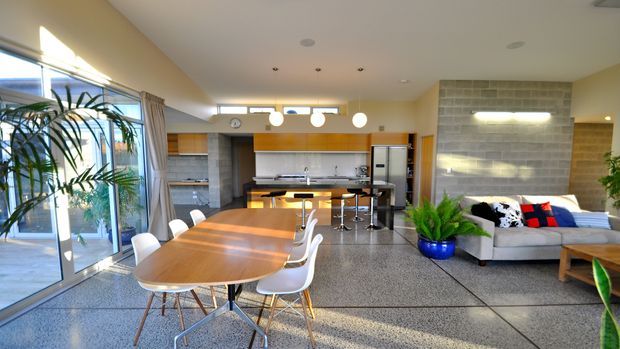 A bright open-plan interior combining a dining area, kitchen and living room. A large timber dining table with white chairs sits in the foreground, while the kitchen features warm wood cabinetry, a long island with stools and stainless-steel appliances. The living area includes a light sectional sofa against a grey block feature wall. Full-height glazing, polished concrete-style flooring and abundant indoor plants create an airy contemporary atmosphere.