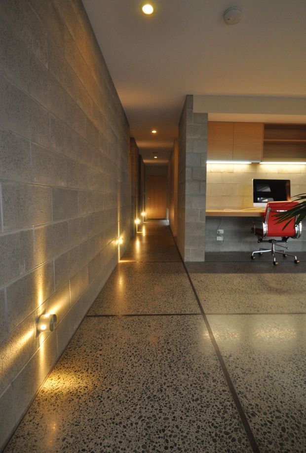 A contemporary interior corridor with exposed grey concrete block walls, polished aggregate concrete flooring, and warm recessed and low-level wall lighting. The hallway opens to a compact home office with timber cabinetry, a desk, monitor, and red office chair.