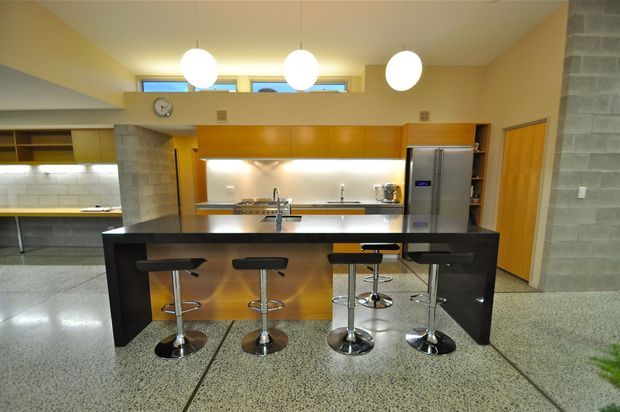 A spacious contemporary kitchen with a large dark island and five bar stools, extensive warm timber cabinetry, stainless-steel appliances, stone benchtops, and a polished terrazzo floor. High clerestory windows and pendant lights provide bright illumination, while exposed concrete block walls add texture.
