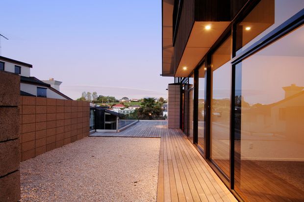 Exterior view of a contemporary residential terrace or deck beside a modern home, with extensive floor-to-ceiling glazing, warm timber soffits and decking, dark metal window frames, and a gravel-covered outdoor area. The terrace overlooks neighbouring suburban homes and a distant coastal-looking landscape, while reflections reveal a sparsely furnished interior.