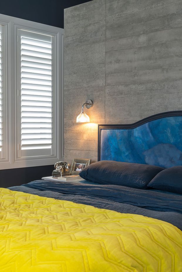 A styled bedroom featuring a dark navy wall, a large grey textured concrete-look feature wall, white plantation shutters, and a bed with a dramatic deep-blue upholstered headboard. Navy bedding and a bright yellow geometric quilt create a bold, high-contrast colour scheme. A wall-mounted bedside light, small bedside table, framed photograph, and decorative accessories complete the room.