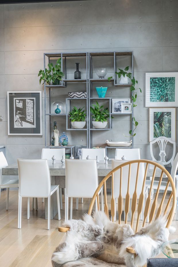 Bright contemporary dining area with a long pale stone dining table, white upholstered dining chairs, and a warm timber spindle-back chair in the foreground. The room features a grey concrete-look paneled feature wall, open metal display shelving with plants and decorative objects, framed botanical and graphic artwork, and light timber flooring. The styling blends clean modern lines with natural greenery and warm wood accents.