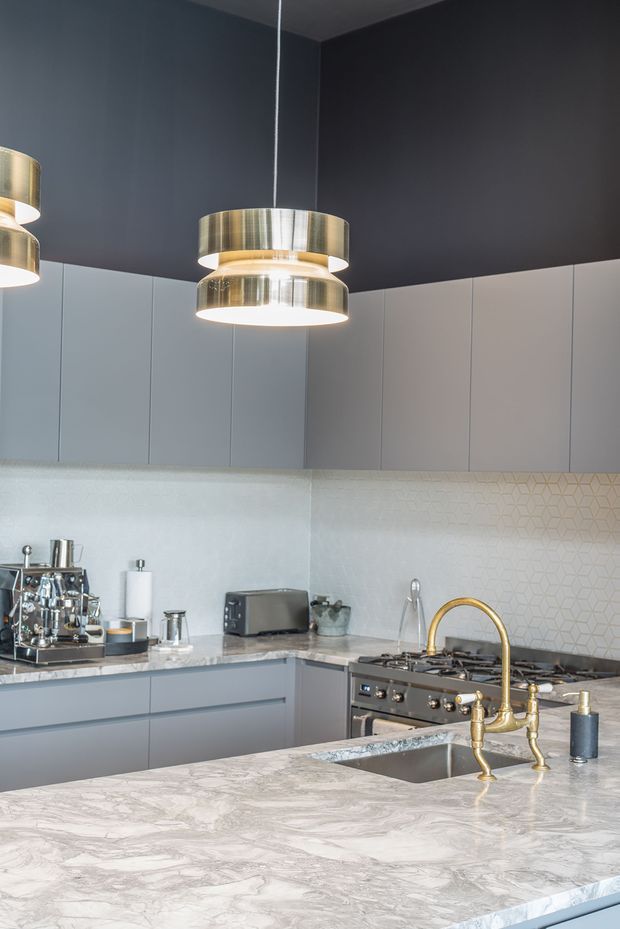 A refined contemporary kitchen with a large marble-look island, integrated grey cabinetry, pale geometric tile splashback, brass tapware and hardware, stainless steel appliances, and layered metallic pendant lighting. The dark charcoal ceiling and upper wall create contrast with the light work surfaces and cabinetry.