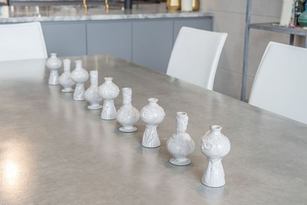 Close-up view of a contemporary dining area dominated by a large smooth grey dining table, with white upholstered dining chairs and a row of decorative white ceramic vases. Grey built-in cabinetry and shelving are visible in the background.
