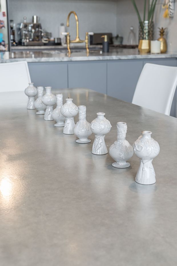 A contemporary kitchen-dining space viewed across a large grey stone or concrete-look dining table. A row of ornate white ceramic candlestick ornaments runs along the table, with white dining chairs in the foreground and background. Behind it is a streamlined grey kitchen with flat-panel cabinetry, a dark marbled benchtop or splashback, a brass gooseneck tap, stainless coffee equipment, and decorative gold-toned vessels. The scene is bright, neutral, and refined, with some greenery and decorative objects.