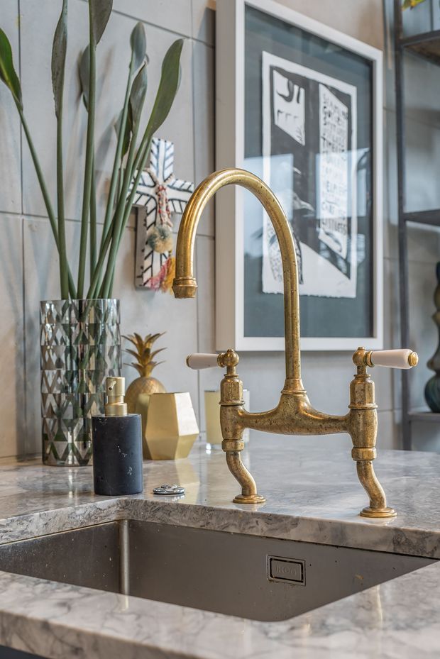 Close-up interior detail of a kitchen sink set into a veined marble countertop, featuring a vintage-style aged brass bridge faucet with porcelain lever handles. A dark undermount sink, soap dispenser, patterned glass vase with tall foliage, decorative objects, framed artwork, and dark-framed glazing are visible. The composition feels refined and eclectic, with warm brass contrasting against cool grey stone and dark accents.