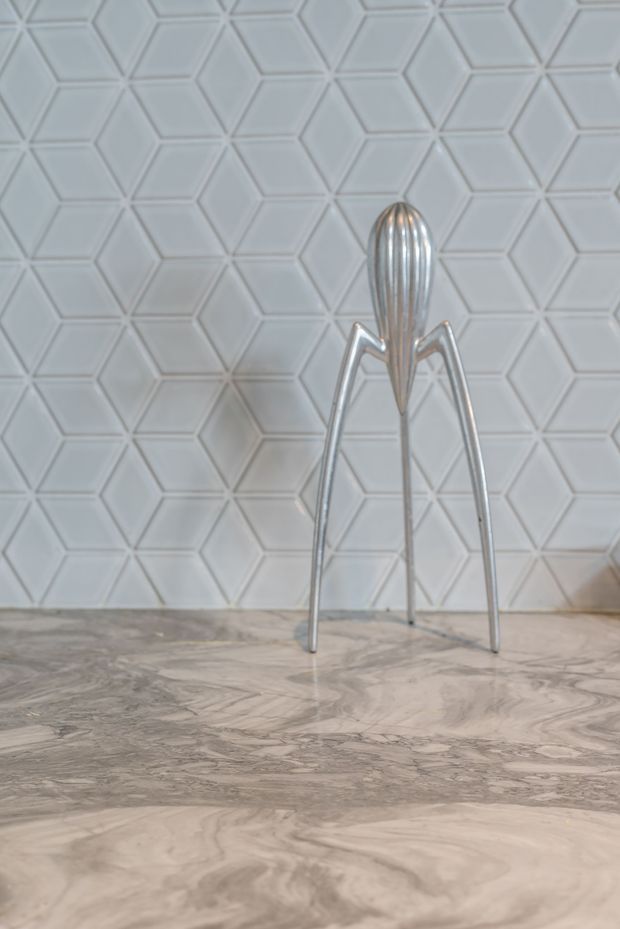 Close-up of a sculptural silver metal citrus juicer standing on a grey-and-white veined stone benchtop, set against a pale embossed geometric hexagonal tile splashback. The image is a product-style detail shot focused on the countertop and wall finish.
