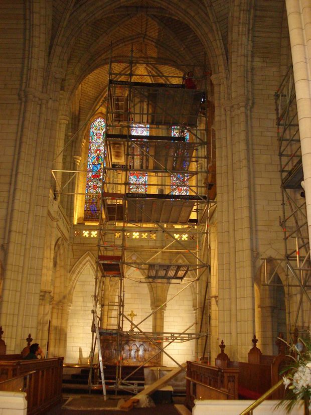 Interior view of a historic Gothic-style church or cathedral undergoing restoration, dominated by tall pale stone masonry walls, pointed arches, ribbed vaulted ceiling, and a large stained-glass window. Extensive metal scaffolding rises through the nave in front of the window and architectural detailing. Dark timber choir or pew railings are visible in the foreground, with warm amber illumination.
