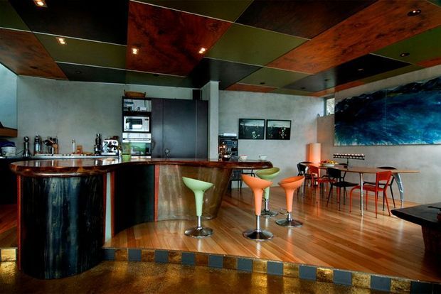 A dark, richly finished open-plan bar and dining space with a curved timber-fronted bar, stainless-steel cylindrical counter, colourful modern bar stools, and a small dining setting. The room features timber flooring, concrete-look walls, dramatic large-format artwork, and a highly distinctive patchwork ceiling of timber, dark panels, and green panels with recessed downlights.