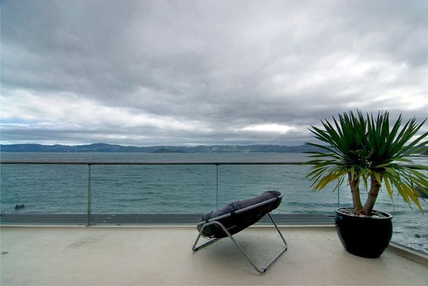 A minimalist waterfront balcony or terrace overlooking calm water and distant hills. The space has a pale concrete or stone deck, frameless glass balustrading with dark metal supports, a reclining outdoor chair, and a large potted tropical plant. The overcast sky and muted blue-grey palette create a serene, contemporary outdoor setting.