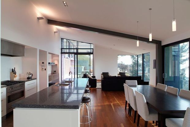 Open-plan contemporary kitchen, dining, and living area with extensive glazing and a double-height-feeling ceiling. The kitchen has sleek white fixed cabinetry, stainless-steel appliances, and a dark polished stone island benchtop with bar stools. Dark timber flooring runs through the space, while a long dark structural beam and black-framed windows and doors add contrast. A dining table and living seating occupy the bright, connected rear portion of the room.