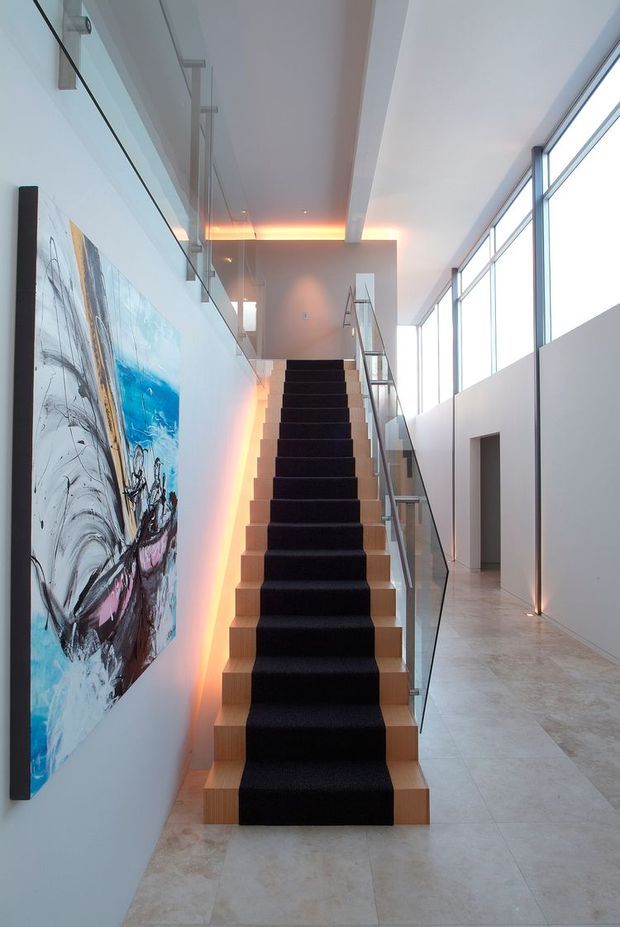 A contemporary interior stairway with a straight timber staircase and dark carpet runner, enclosed by clear glass balustrades. The stair rises through a bright, double-height circulation space with full-height windows, pale stone flooring, white walls, recessed ceiling details and warm concealed LED lighting. A large abstract artwork forms a feature on the left wall.