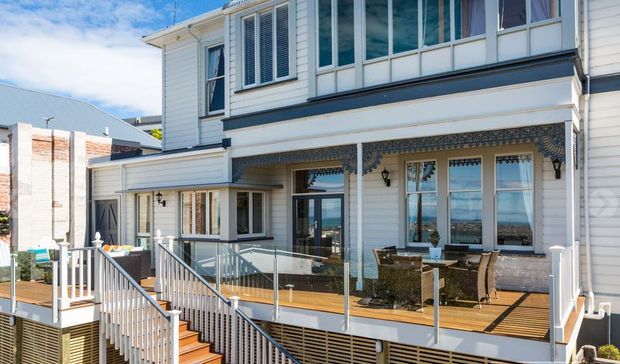 Exterior view of a multi-level coastal-style home with white weatherboard cladding, extensive glazing, decorative patterned tile detailing beneath the upper balcony, and a timber deck with glass balustrades and stairs. The architecture overlooks a bright blue coastal or waterfront setting visible through the openings.
