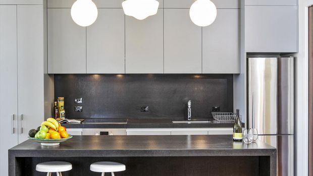 Contemporary compact kitchen with a dark stone-look island and matching full-height splashback, pale grey flat-panel cabinetry, stainless-steel refrigerator and appliances, and three white pendant lights. The island includes seating for two, with a fruit bowl and small accessories on the benchtop.