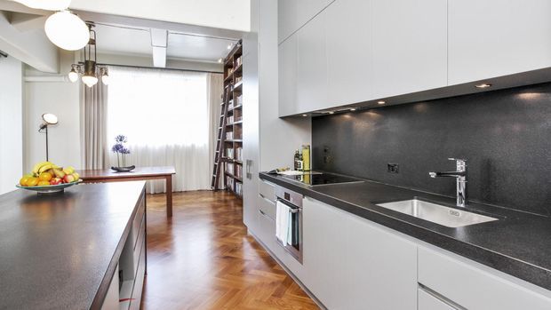 A bright contemporary kitchen with a long dark stone benchtop, integrated stainless-steel sink and appliances, and sleek white handleless cabinetry. A central island opens toward a dining area with a timber table, pendant lights, bookshelves, and parquet flooring. Large sheer-curtained glazing brings in soft natural light.