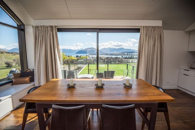 A bright dining area centered on a substantial warm timber dining table with dark upholstered chairs. Full-height glazing and drawn curtains open to a panoramic view of a green sports field, lake, and mountains. A compact white built-in kitchen or cabinetry zone is visible at right, while dark timber flooring, neutral walls, and a paneled ceiling complete the room.