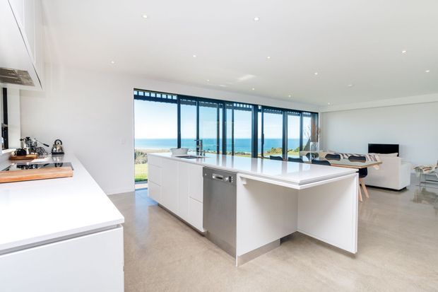 Bright, open-plan contemporary kitchen with a large white island and integrated seating, positioned in front of full-height glazed doors overlooking a blue coastal/ocean view. White cabinetry and benchtops, stainless-steel dishwasher, polished concrete-look flooring, recessed ceiling lights, and a connected living area are visible.