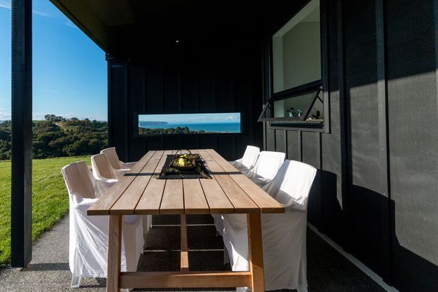 Covered outdoor dining area beside a dark-clad building, with a long timber dining table surrounded by white slipcovered chairs. The open side frames a sunny green landscape and distant coastal-looking horizon, while a high awning window and large opening provide ventilation and views. The space feels relaxed, contemporary, and connected to the outdoors.