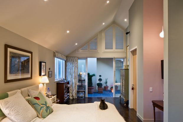 A warm, traditionally styled bedroom viewed toward a bright glazed sitting or courtyard area. The room has a pitched, vaulted ceiling with recessed spotlights, cream walls, dark timber flooring, a large bed with pale bedding and decorative cushions, bedside lighting, framed artwork, and extensive upper glazing fitted with white plantation shutters. A small cat rests on the bed. Beyond the central glazing is a tiled outdoor or sunroom-like area with potted plants.