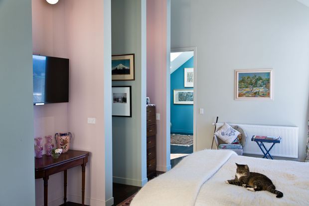A colorful bedroom viewed from the sleeping area, featuring a neatly made bed with a cat, pale blue-grey walls, a pink-painted structural partition, wall-mounted television, dark timber console, framed artwork, radiator, and an open doorway leading to a vivid turquoise adjoining space. The room has traditional detailing combined with playful, eclectic colour and furnishings.