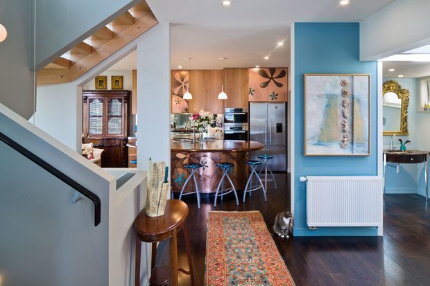 A bright, eclectic open-plan interior viewed from a stairway, with a kitchen and dining area ahead. The room features warm timber cabinetry and flooring, a central island with curved timber seating, colorful patterned artwork, a blue painted feature wall, recessed lighting, and a glimpse of a bathroom or vanity area to the right. A timber staircase with dark handrail and balustrade occupies the foreground.