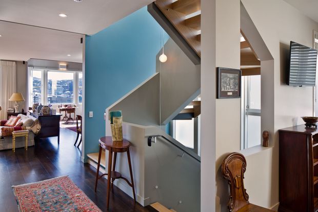 Interior view of a refined multi-level home showing an entry and stairway area opening into a living room. The staircase has warm timber treads and open risers, a grey painted side wall, and a vivid blue feature wall. Dark timber flooring continues through the circulation space and living area, with classic furnishings, framed artwork, a pendant light, and a wall-mounted television visible.
