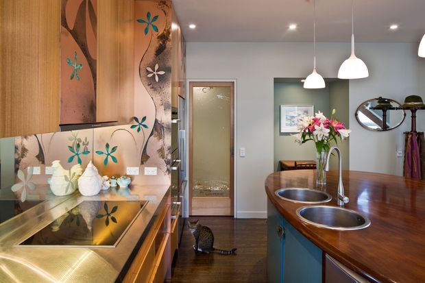 A warm, eclectic kitchen and dining space viewed from the kitchen work area. The foreground features timber cabinetry, a reflective patterned floral backsplash, a dark reflective benchtop, and an integrated cooktop. A timber island with two round stainless-steel sinks and a curved faucet separates the kitchen from the dining area. Pendant lights hang above the island, while the adjoining room has muted green walls, timber flooring, a framed artwork, a round wall mirror, and a timber dining table. Decorative floral motifs and sculptural objects add a playful, artistic character.