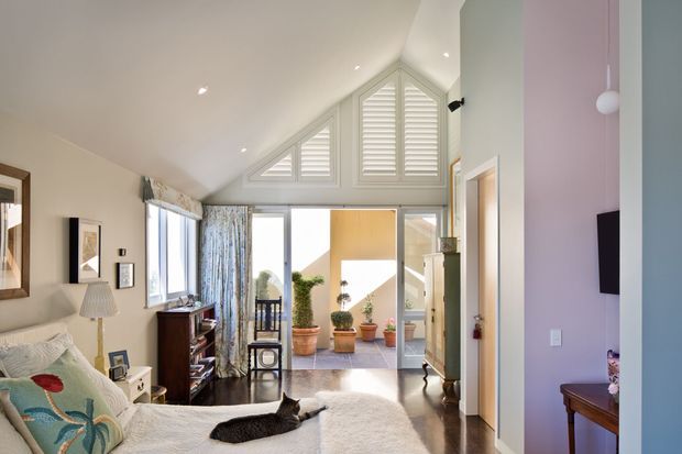 Bright open-plan living area with a sloped ceiling and prominent triangular clerestory windows fitted with white plantation shutters. A bedroom or sleeping area with a bed, bedside table, lamp and cat occupies the foreground, while the space opens toward a glazed courtyard or sunlit outdoor area with potted plants. Dark polished timber flooring, pale walls, framed artwork and a compact timber console contribute to the warm, relaxed interior.