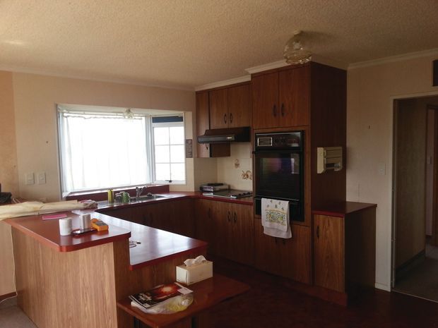 A compact traditional kitchen with dark timber cabinetry, reddish laminate benchtops, integrated appliances, a peninsula, and a small adjoining dining area. The room has warm beige walls, dark flooring, and a large window providing natural light.