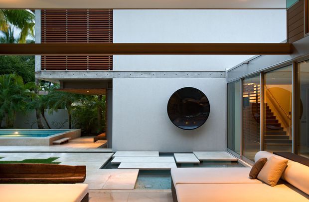 A sophisticated modern courtyard viewed from an outdoor lounge area, featuring a shallow reflective water feature with stepping stones, pale stone paving, lush tropical planting, a smooth rendered facade with a circular window, extensive glazing, timber privacy screens, and a warmly lit stairway. Built-in outdoor seating and a sculptural timber bench frame the foreground.