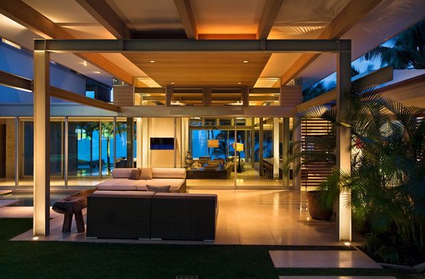 Evening view from an outdoor lawn or terrace into a sophisticated open-plan living space. The home features extensive floor-to-ceiling glazing, warm timber ceilings and beams, slim dark metal framing, polished light flooring, layered indoor and outdoor seating, and lush tropical planting. Dramatic integrated lighting creates a warm, luxurious atmosphere, while the visible upper level and strong horizontal and vertical framing suggest a contemporary multi-level residence.