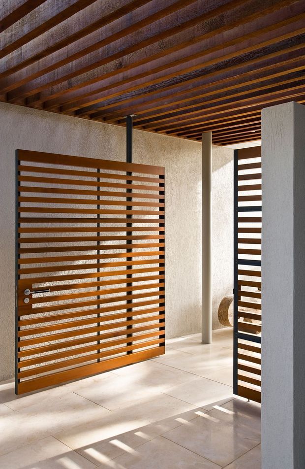 A bright, minimalist covered entry courtyard with white textured plaster walls, pale large-format tiled flooring, and strong horizontal timber slatted screens and gate set in dark metal frames. The ceiling is composed of exposed dark timber rafters, while slender round columns support the sheltered structure. Sunlight casts crisp linear shadows across the floor, creating a warm, contemporary architectural composition.