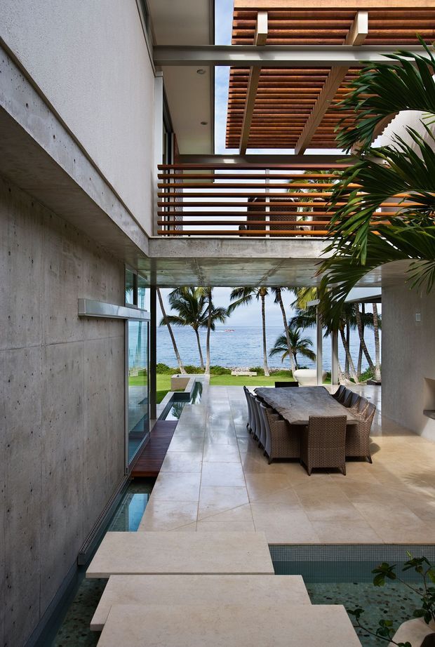 A modern tropical coastal outdoor dining courtyard beside the ocean. The open-air space features a long outdoor dining table with woven chairs, pale stone paving, exposed concrete walls and soffits, large glass openings, timber slatted pergola and privacy screens, tropical palms, and a small reflecting pool or water feature along the foreground. The composition looks through the courtyard toward a beachfront and ocean.
