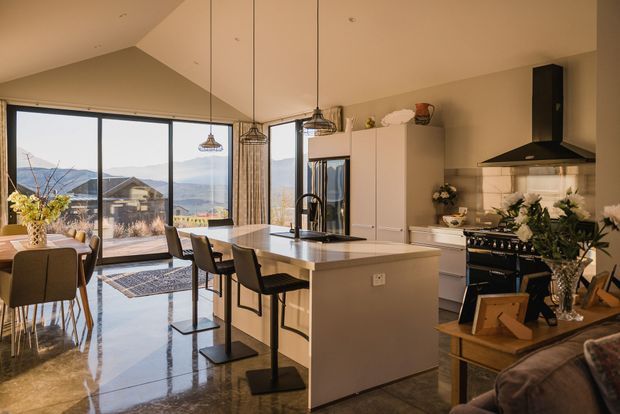 A bright, open-plan kitchen and dining area with a large central island, white cabinetry, black pendant lights, and a freestanding range beneath a dark sculptural rangehood. Floor-to-ceiling glazing opens to a terrace and distant mountainous landscape. The polished dark floor reflects the warm natural light, while a dining table and bar seating define the space.