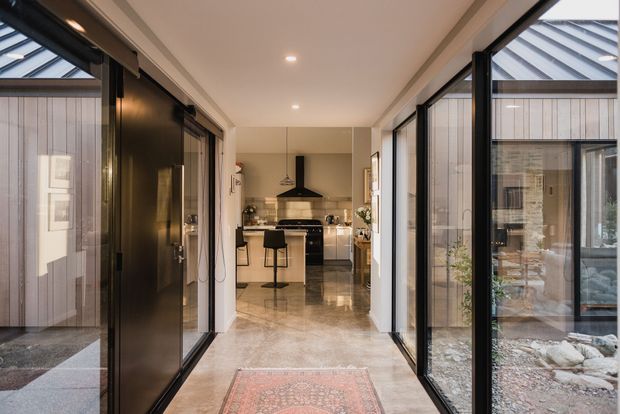 A bright, narrow glazed transition space or hallway leading directly into a contemporary kitchen. Black-framed full-height glazing and doors line both sides, with polished stone-look flooring, a patterned runner, recessed ceiling lights, and views into an adjacent courtyard and entry area. The kitchen beyond includes a central island with bar seating, dark cabinetry, a rangehood, and light stone surfaces.