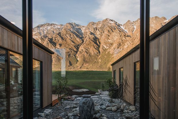 A view through a narrow landscaped courtyard between two dark timber-clad wings, with full-height black-framed glazing and a central gravel-and-stone garden featuring a sculptural water vessel. Dramatic rugged mountain peaks and a grassy alpine landscape rise directly beyond the property. The composition emphasises a strong indoor–outdoor connection, natural materials, and a sheltered mountain setting.
