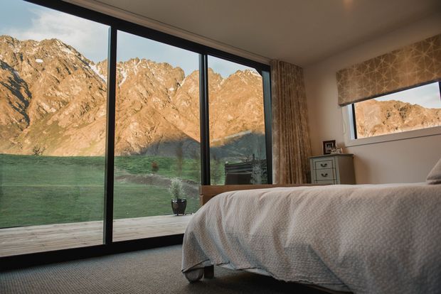 A serene bedroom with a large bed facing expansive floor-to-ceiling black-framed glazing. The windows open to a timber deck and dramatic rugged mountain scenery, while a smaller window, patterned Roman blind, curtains, and a compact bedside chest complete the warm, softly styled interior.