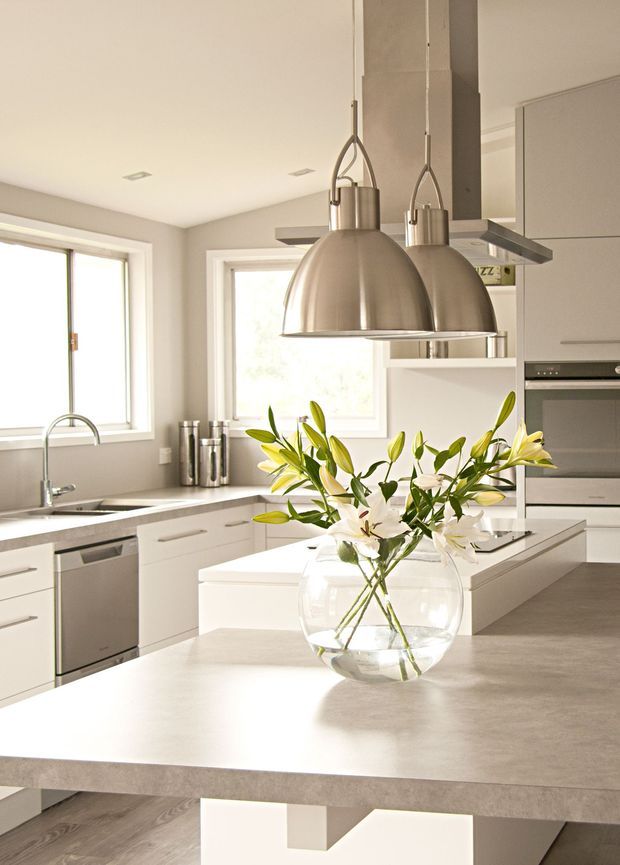 Bright contemporary kitchen with white flat-panel cabinetry, stainless-steel appliances, a large grey island benchtop, double stainless-steel pendant lights, and broad windows providing natural light. A glass vase of lilies sits on the island.