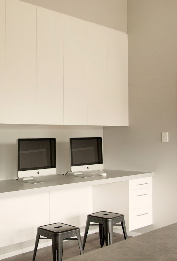 Minimalist home office with two desktop computers positioned on a long grey worktop, extensive white built-in overhead storage, and matching white base cabinetry with drawers. Two black metal stools sit at the workstation, which is set against a warm light-grey wall with carpeted flooring.