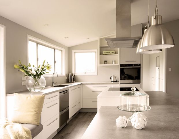 Bright contemporary kitchen with a white L-shaped cabinetry layout, central island, grey benchtops, integrated stainless-steel appliances, and large windows. A sculptural pendant light hangs above the island, while a small upholstered seating area and vase of greenery soften the space.