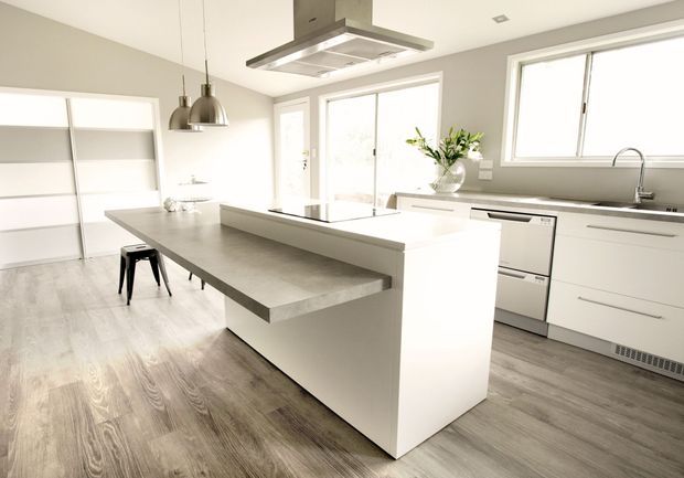 Bright, minimalist contemporary kitchen with a large central island, white handleless cabinetry, a grey concrete-look benchtop, integrated stainless-steel rangehood, pendant lights, and grey-toned timber flooring. Large windows and glazed doors provide abundant natural light, while open shelving and a small stool occupy the adjoining area.