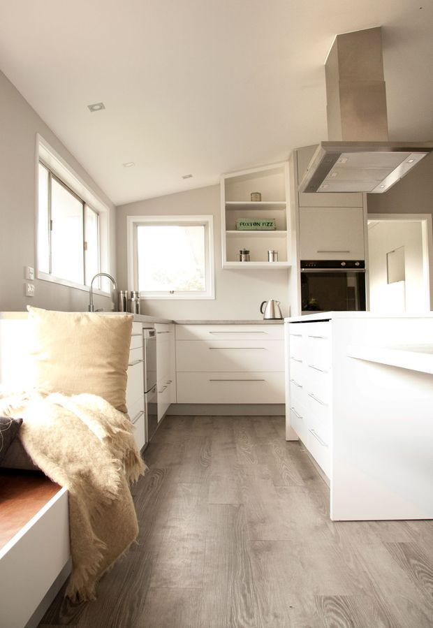 Bright, compact contemporary kitchen with white handleless cabinetry, a large central island, integrated oven, stainless-steel rangehood, pale stone benchtops, grey timber-look flooring, open shelving, and generous windows. A relaxed seating nook with a cream throw is visible in the foreground.