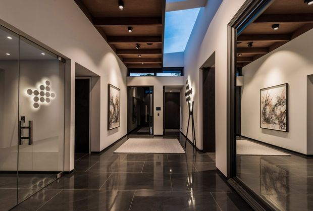 A refined modern hallway or entry corridor with glossy dark stone tile flooring, smooth light walls, dark timber ceiling panels with recessed spotlights, and a dramatic linear skylight. Full-height mirrored or glazed surfaces reflect the corridor, artwork, and sculptural wall lighting. The space has dark interior doors, minimal black trims, and a pale rectangular runner, creating a sophisticated, gallery-like atmosphere.