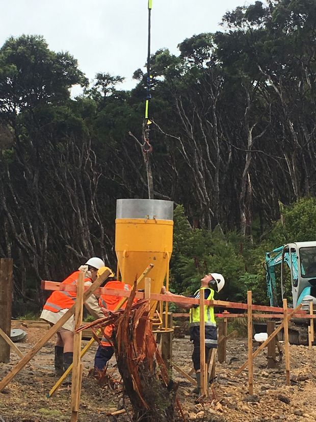 Outdoor construction site in a wooded setting, with workers wearing high-visibility safety clothing and hard hats. A large yellow concrete or aggregate hopper is suspended above the work area, with construction equipment and temporary timber barriers visible.