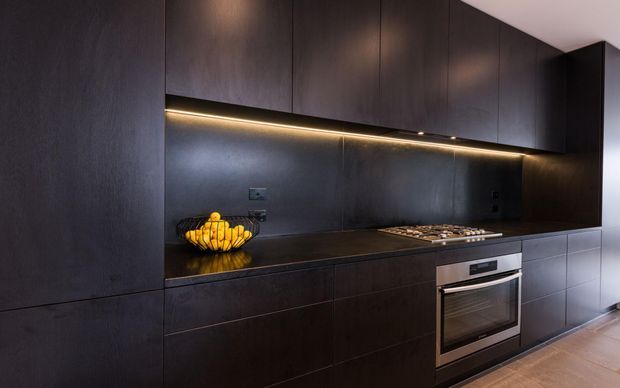 A sleek, dark-toned contemporary kitchen with full-height integrated cabinetry, a continuous dark benchtop and matching splashback, under-cabinet LED strip lighting, and a built-in stainless-steel oven and cooktop. The minimalist joinery creates a moody, refined atmosphere, with warm timber flooring adding contrast.