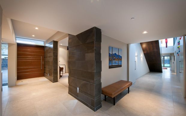 Contemporary residential entry hall with a large timber pivot front door, warm stone-clad structural columns, pale tiled flooring, recessed ceiling lights, a leather bench, and artwork. The corridor extends toward a brighter living area with skylight-like glazing and additional built-in elements.